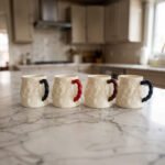 Four off-white textured ceramic coffee mugs with contrasting black, red, brown, and blue handles lined up on a marble kitchen countertop.