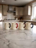 Four off-white textured ceramic coffee mugs with contrasting black, red, brown, and blue handles lined up on a marble kitchen countertop.