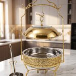Luxury gold and stainless steel round chafing dish buffet server with a glass dome lid suspended on a gold frame, matching ladle, and buffet burner on a kitchen counter.
