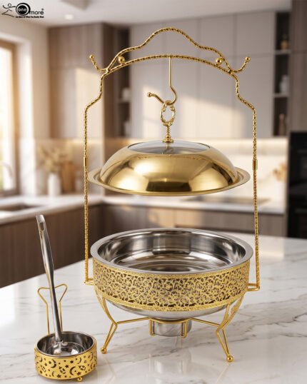 Luxury gold and stainless steel round chafing dish buffet server with a glass dome lid suspended on a gold frame, matching ladle, and buffet burner on a kitchen counter.