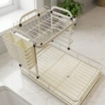 Cream two-tier metal dish drying rack with side utensil holder and drain tray on a kitchen countertop next to a sink.