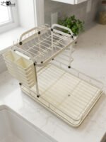 Cream two-tier metal dish drying rack with side utensil holder and drain tray on a kitchen countertop next to a sink.