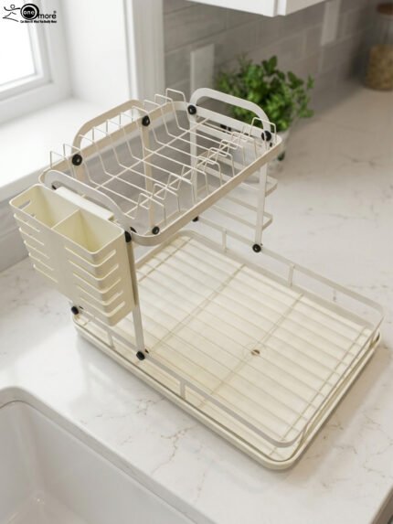 Cream two-tier metal dish drying rack with side utensil holder and drain tray on a kitchen countertop next to a sink.
