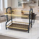 Two-tier gold and black metal dish drying rack with removable drip tray and utensil holder on a white marble kitchen countertop.
