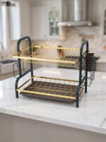 Two-tier gold and black metal dish drying rack with removable drip tray and utensil holder on a white marble kitchen countertop.