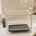 White 2-tier metal wire dish drying rack with removable dark gray drain tray and side utensil holder on a marble kitchen countertop.
