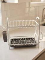 White 2-tier metal wire dish drying rack with removable dark gray drain tray and side utensil holder on a marble kitchen countertop.