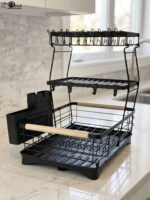 3-tier black wire dish drying rack on a marble countertop with wooden handles, attached utensil holder, and removable drip tray