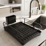 Matte black metal dish drying rack with attached utensil holder and drip tray sitting on a marble kitchen countertop beside a sink.