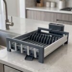 Modern grey and silver expandable kitchen dish drying rack with plate slots, utensil caddy, and drain spout sitting on a countertop next to a sink.