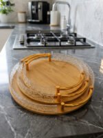 Set of three round wooden nesting serving trays with curved gold handles and clear textured glass rims stacked on a grey marble kitchen countertop.