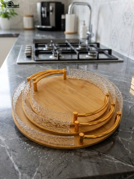 Set of three round wooden nesting serving trays with curved gold handles and clear textured glass rims stacked on a grey marble kitchen countertop.