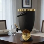 Matte black 5.5L beverage cooler dispenser with gold metallic accents, top handle, and dispensing tap resting on a dining table