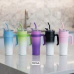 Five 1180ml ombre stainless steel tumblers with handles and straws in blue, green, purple, black, and pink lined up on a marble kitchen counter.