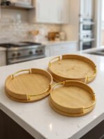Set of 3 nesting round bamboo wooden serving trays with stylish gold metal handles on a white countertop.