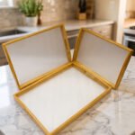 Set of three natural wood nesting serving trays with cutout handles and smooth white inner bases displayed on a kitchen marble countertop.