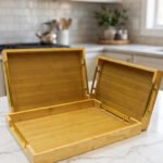 Set of three rectangular natural wood serving trays with gold metal handles displayed on a white marble kitchen countertop.