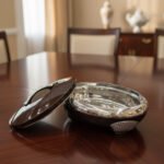 An elegant oval-shaped insulated hotpot in dark brown with a silver stainless steel interior and decorative studded handles, open with the lid resting on a polished wooden dining table.