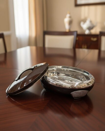 An elegant oval-shaped insulated hotpot in dark brown with a silver stainless steel interior and decorative studded handles, open with the lid resting on a polished wooden dining table.