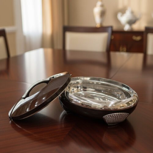An elegant oval-shaped insulated hotpot in dark brown with a silver stainless steel interior and decorative studded handles, open with the lid resting on a polished wooden dining table.