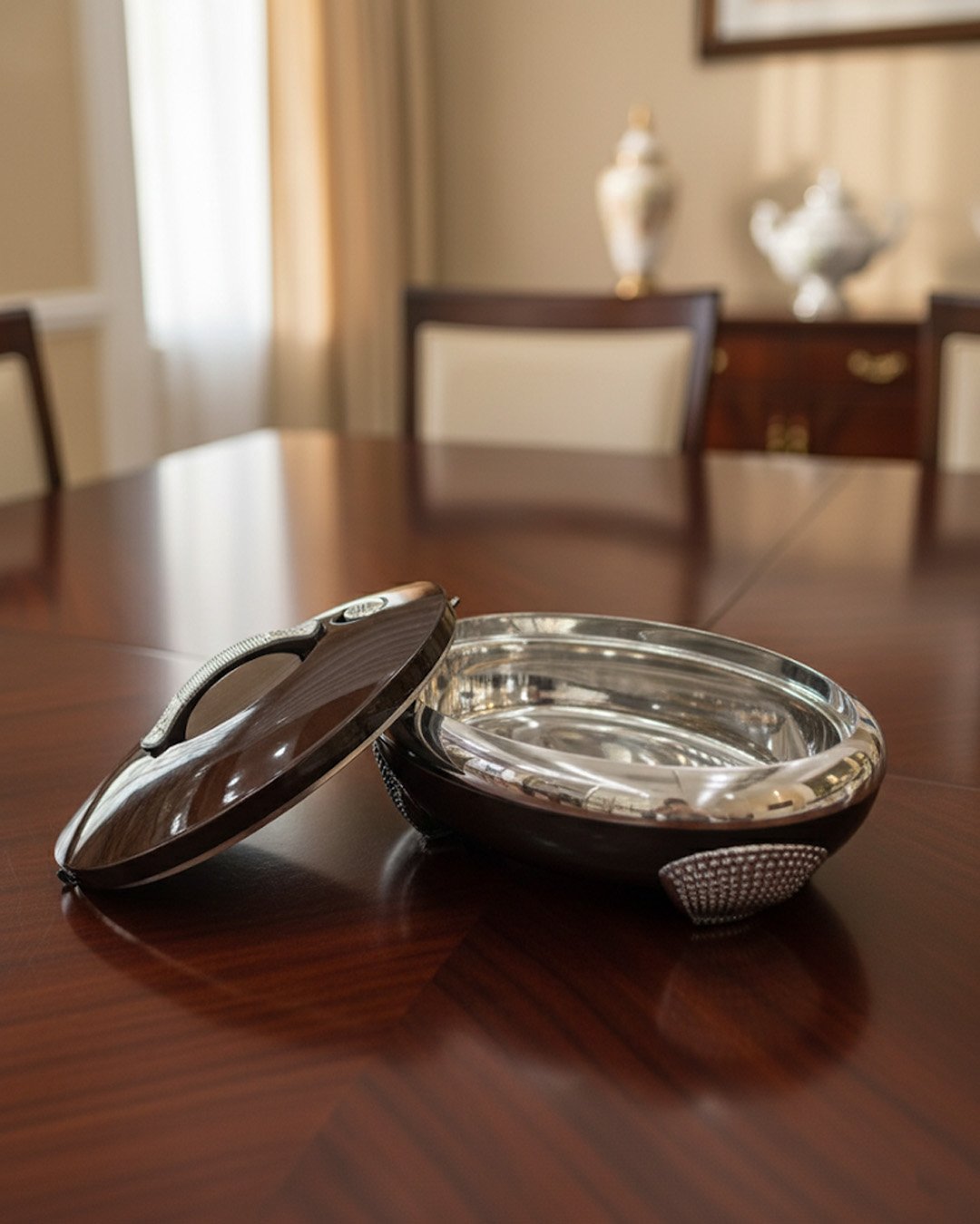 1 An elegant oval-shaped insulated hotpot in dark brown with a silver stainless steel interior and decorative studded handles, open with the lid resting on a polished wooden dining table.