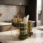 Luxury emerald green ceramic soup tureen set with gold accents, glass lid, warmer stand, stacked double-handled cups, saucers, and white spoons on a marble countertop.