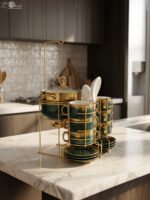 Luxury emerald green ceramic soup tureen set with gold accents, glass lid, warmer stand, stacked double-handled cups, saucers, and white spoons on a marble countertop.