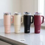 Four 1180ml insulated stainless steel tumblers with handles and clear lids in pink, cream, black, and burgundy lined up on a kitchen counter.