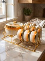 Ceramic soup tureen set with orange gradient rims and gold accents, including hanging soup cups, saucers, and spoons organized on a gold metal rack.