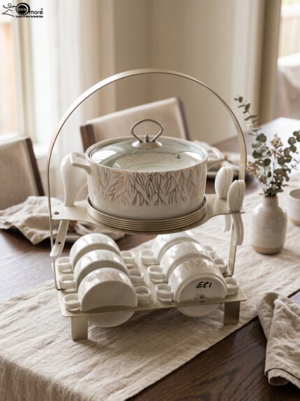 Ceramic soup tureen with clear glass lid, matching soup bowls with handles, saucers, and spoons arranged on a two-tier metal stand on a wooden dining table.