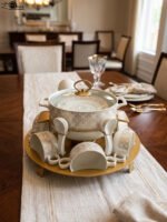 Luxury white and gold ceramic soup tureen set with warming stand, matching bowls, saucers, spoons, and glass lid on a wooden table.