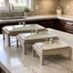 Three rectangular ceramic single buffet dishes with clear glass lids set in champagne gold stands on a kitchen marble countertop.