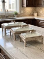 Three rectangular ceramic single buffet dishes with clear glass lids set in champagne gold stands on a kitchen marble countertop.