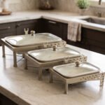 Three ceramic single buffet dishes with clear glass lids and champagne gold stainless steel warming stands displayed on a marble kitchen counter.