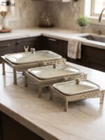 Three ceramic single buffet dishes with clear glass lids and champagne gold stainless steel warming stands displayed on a marble kitchen counter.