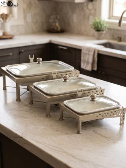 Three ceramic single buffet dishes with clear glass lids and champagne gold stainless steel warming stands displayed on a marble kitchen counter.