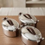 Set of three One More polished stainless steel oval hot pots with brown leather handles in 2L, 2.5L, and 3L sizes on a wooden tabletop.