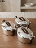 Set of three One More polished stainless steel oval hot pots with brown leather handles in 2L, 2.5L, and 3L sizes on a wooden tabletop.