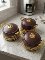 Set of three deep maroon insulated hot pots with gold handles and honeycomb gold accent trim on a kitchen counter.