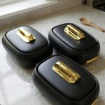 Three matte black insulated hot pot food containers with gold lid handles resting on a marble kitchen countertop.