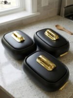 Three matte black insulated hot pot food containers with gold lid handles resting on a marble kitchen countertop.
