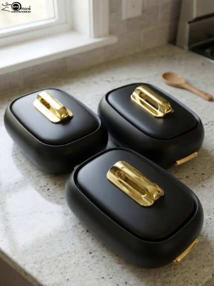 Three matte black insulated hot pot food containers with gold lid handles resting on a marble kitchen countertop.