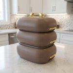 Three stacked matte taupe One More insulated hot pots with gold handles on a white kitchen countertop.