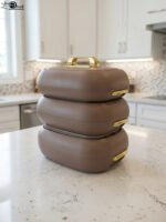 Three stacked matte taupe One More insulated hot pots with gold handles on a white kitchen countertop.