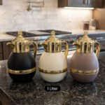 Three luxury 1-liter thermal coffee and tea vacuum flasks with ornate gold lids and accents in matte black, cream, and taupe on a granite kitchen countertop.