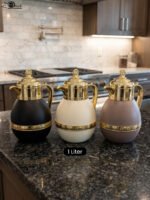 Three luxury 1-liter thermal coffee and tea vacuum flasks with ornate gold lids and accents in matte black, cream, and taupe on a granite kitchen countertop.