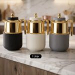 Three One More 1-liter luxury thermal vacuum flasks with polished gold-tone tops and textured ribbed bases in matte black, off-white, and grey resting on a marble kitchen countertop.