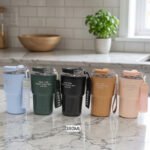Set of 380ml portable travel coffee mugs in light blue, dark green, matte black, camel, and pink with attached wrist straps on a marble countertop.