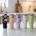 Five 1180ml floral stainless steel travel tumblers with handles in black, peach, lavender, white, and sage green displayed on a kitchen counter.