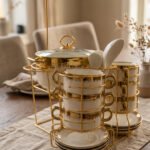 White ceramic soup tureen and stacked double-handled soup bowls with gold rim accents on a gold wire stand on a dining table.